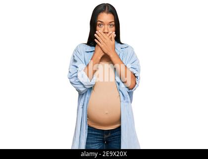 Belle femme hispanique s'attendant à un bébé montrant enceinte ventre choqué couvrant la bouche avec des mains pour erreur. Concept secret. Banque D'Images