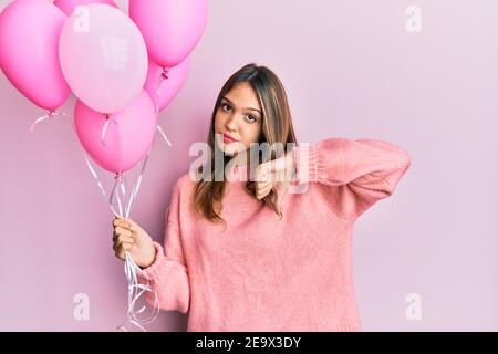 Jeune femme brunette tenant des ballons roses avec le visage en colère, signe négatif montrant le désaccord avec les pouces vers le bas, concept de rejet Banque D'Images