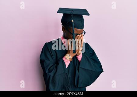 Jeune fille afro-américaine portant une casquette de remise des diplômes et une robe de cérémonie avec une triste expression couvrant le visage avec les mains en pleurant. Concept de dépression. Banque D'Images