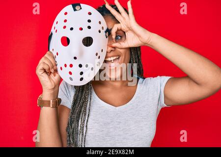 Jeune femme afro-américaine aux tresses portant un masque de hockey souriant bonne marche ok avec la main sur l'oeil à travers doigts Banque D'Images