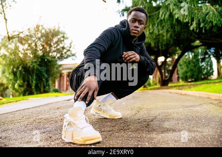Petit angle de jeune homme ethnique dans des vêtements élégants de squating sur le chemin entre les arbres et regardant la caméra en ville Banque D'Images