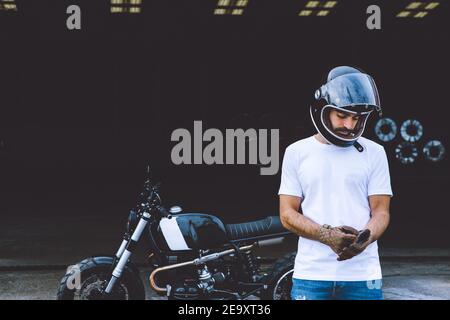 Corps complet d'un jeune homme de race sérieuse dans une ambiance décontractée vêtements et casquette à proximité de la moto, près des grues industrielles à bord de mer Banque D'Images