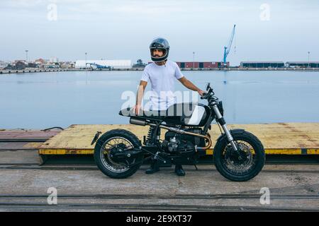 Jeune homme ethnique à barbe confiant en t-shirt blanc et jeans en moto debout au bord de la mer Banque D'Images