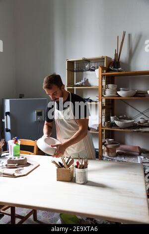 potier masculin barbu jeune concentré dans des vêtements et un tablier décontractés création d'une plaque en céramique blanche en studio Banque D'Images