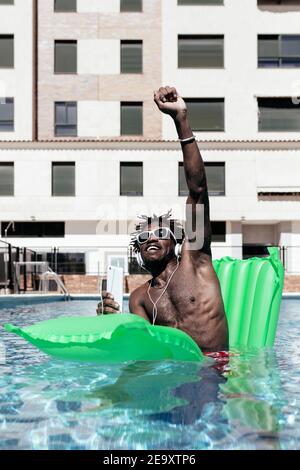 Ravi Afro-américain homme assis sur un matelas gonflable dans la natation profitez de la piscine et écoutez de la musique dans les écouteurs tout en profitant de l'été vacances wi Banque D'Images