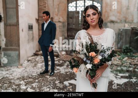 Contenu jeune mariée portant robe de mariage élégante avec bouquet tendre en regardant la caméra près d'un marié ethnique dans obsolète ruiné bâtiment Banque D'Images