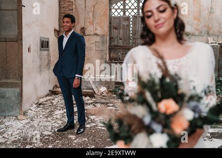 Contenu jeune mariée portant robe de mariage élégante avec bouquet tendre se tenir près du marié ethnique dans une salle de bâtiment obsolète en ruines Banque D'Images