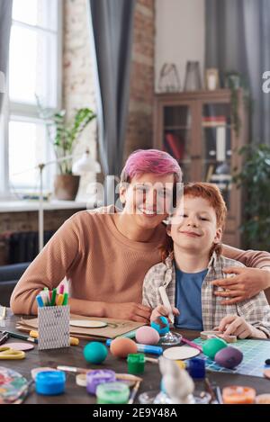 Portrait de mère et de fils souriant à l'appareil photo tout en peignant Œufs à la table et Pâques à la maison Banque D'Images