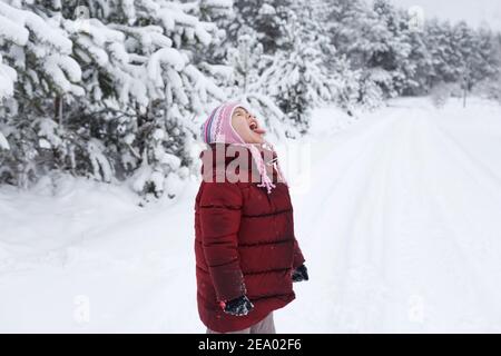 une petite fille dans une veste rouge se tient dans une forêt enneigée. Jeux pour enfants dans la forêt enneigée. Vacances d'hiver en famille avec un enfant Banque D'Images