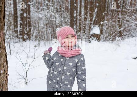 une petite fille se tient dans une forêt enneigée et regarde vers le haut. Jeux pour enfants dans la forêt d'hiver. Vacances d'hiver en famille avec un enfant Banque D'Images