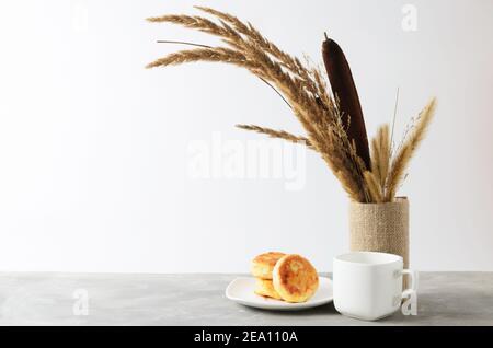 Quelques crêpes sur une assiette blanche et une tasse vide sur une table grise texturée contre un mur blanc avec un bouquet d'automne d'herbes séchées et de roseaux. Sélectionnez Banque D'Images
