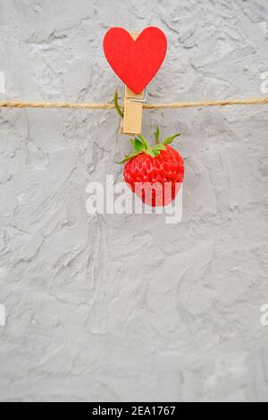 Une fraise mûre rouge accrochée à une épinglette en bois avec des coeurs sur une ficelle de corde, située sur le bord supérieur sur fond de pierre grise. Banque D'Images
