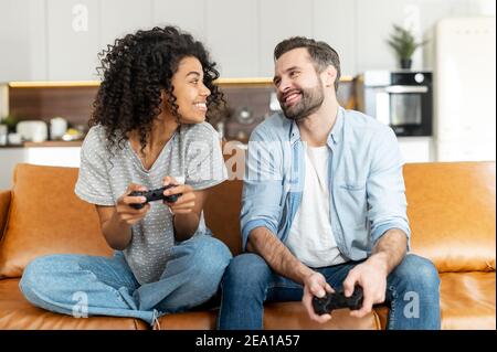 Un couple multiracial excité aime jouer au jeu vidéo, une femme africaine et un homme caucasien tiennent des joysticks sans fil se regarder les uns les autres prêts à combattre Banque D'Images