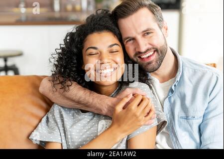 Charmant couple multiracial dans embrasse dans le salon confortable à la maison. Un homme caucasien souriant embrasse une charmante fille africaine. Amour et affection Banque D'Images