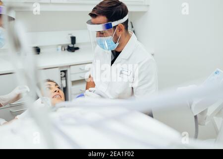 Dentiste traitant une patiente à la clinique dentaire. Un médecin de sexe masculin portant un masque et un écran facial discutant du traitement avec le patient à la clinique dentaire. Banque D'Images