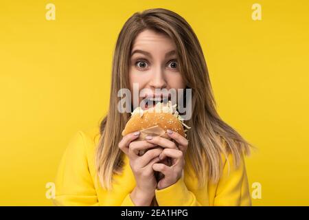 Photo de la belle dame pose d'une manière heureuse essaie de mordre le hamburger. Porte un pull à capuche jaune décontracté, un arrière-plan jaune isolé Banque D'Images