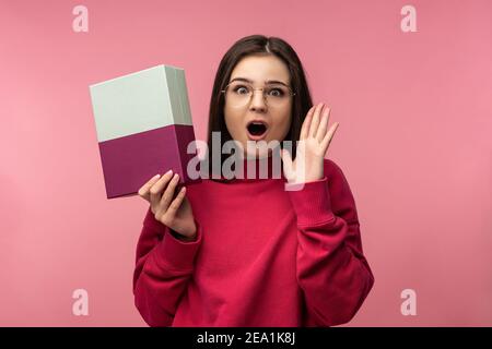 Photo de la belle dame en verres tient boîte avec le présent, les singes surpris. Porte un pull décontracté rose pantalon blanc isolé couleur rose fond Banque D'Images