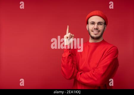 Photo d'un homme attrayant avec une barbe en lunettes et des vêtements rouges. Joli et mignon points mâles vers le haut, isolé sur fond rouge Banque D'Images
