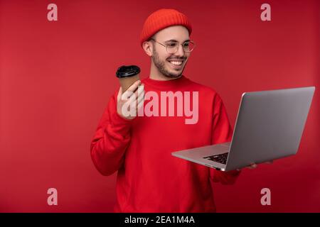 Photo d'un homme attrayant avec une barbe en lunettes et des vêtements rouges. Un homme mignon tient un ordinateur portable et un café, isolé sur fond rouge Banque D'Images