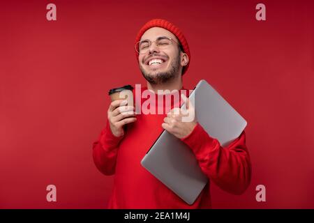 Photo d'un homme attrayant avec une barbe en lunettes et des vêtements rouges. Un homme heureux tient un ordinateur portable et le café se sent bien, isolé sur fond rouge Banque D'Images