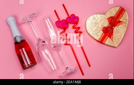 Deux verres à champagne avec pailles à boire en plastique rouge avec coeurs, boîte cadeau et bouteille de champagne rouge sur fond rose. Banque D'Images