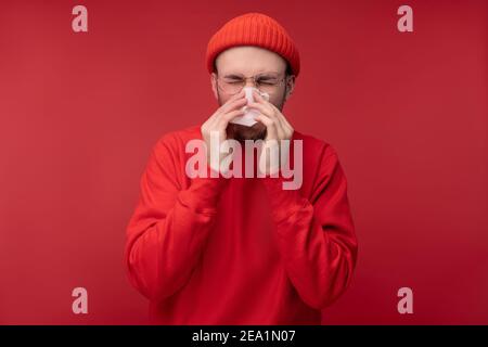 Photo de l'homme heureux avec la barbe dans les vêtements rouges éternuements maladie, isolé sur fond rouge Banque D'Images