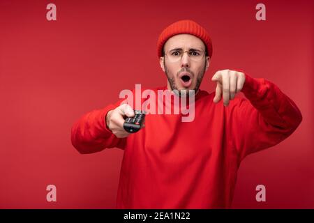 Photo de l'homme agité avec la barbe dans des vêtements rouges pointant les doigts vers l'appareil photo tient la console de télévision avec surprise et la bouche ouverte, isolée sur le rouge Banque D'Images