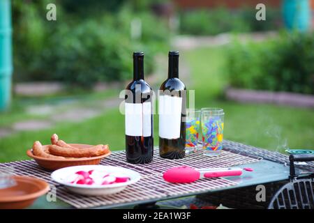 De délicieuses saucisses juteuses du gril sont posées dans une assiette sur une table en osier, sur fond d'herbe verte. Près de deux bouteilles de vin et verres. Banque D'Images