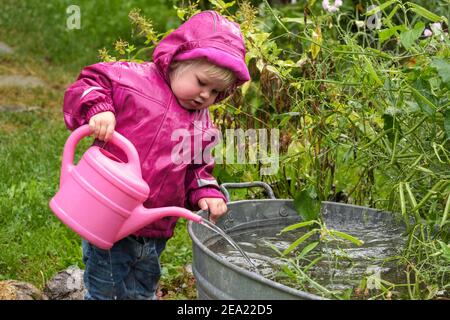 Petite fille avec bottes en caoutchouc et veste de pluie jouant avec l'eau et arrosoir dans la pluie, haute-Bavière, Bavière, Allemagne Banque D'Images