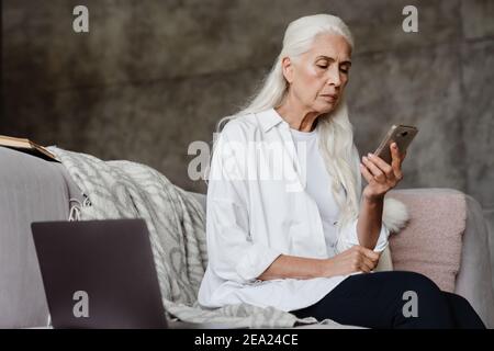 Femme âgée pensive assise sur un canapé à la maison à l'aide d'un téléphone portable et ordinateur portable Banque D'Images