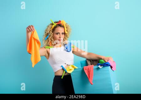 Femme de ménage fatiguée pleine de vêtements et de chiffons à nettoyer. Fond cyan Banque D'Images