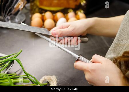 Femme aiguisant couteau de cuisine avec pierre à affûter dans la cuisine, couteau de cuisine. Vue rapprochée des mains. Banque D'Images