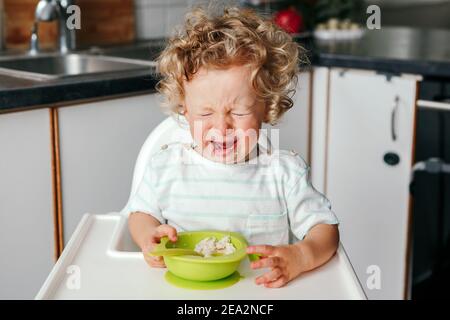 Un jeune garçon caucasien pleure assis sur une chaise haute avec une purée de céréales sur l'assiette. Enfant refusant de manger des aliments sains. Un enfant hurle dans une colère. Deux terribles Banque D'Images