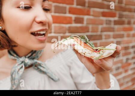 Bonne fille asiatique mangeant un morceau de délicieuse pizza italienne chaude contre un mur de briques vintage. Concept de régime et de gluttonie Banque D'Images