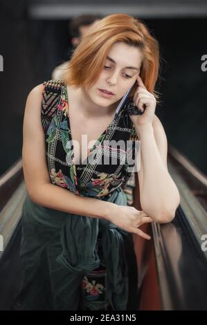 Une jeune femme en dépression parle sur un téléphone intelligent tout en se tenant sur un escalier roulant. Les gens modernes avec des téléphones cellulaires à l'extérieur. Technologie de communication mobile. Haute a Banque D'Images