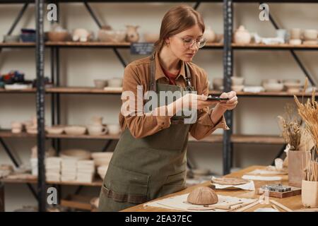 Portrait d'une jeune femme prenant une photo de smartphone d'un bol en céramique brute dans un atelier de poterie à publier sur les médias sociaux, le concept de petite entreprise, l'espace de copie Banque D'Images