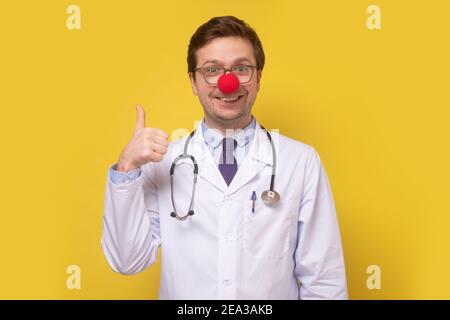 Drôle de docteur caucasien avec le nez rouge clown souriant à la caméra montrant le pouce vers le haut. Prise de vue en studio Banque D'Images