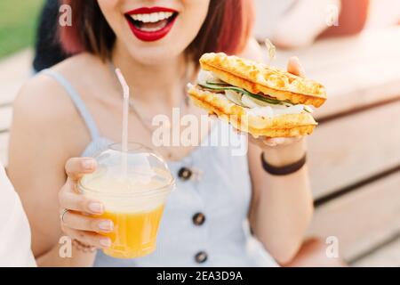 Une femme hipster souriante boit du jus de fruits frais et mange un délicieux sandwich avec un pain de gaufres viennoises à carreaux. Banque D'Images