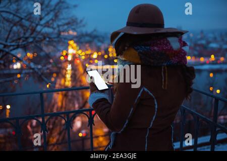 Vue de derrière une femme d'âge moyen dans un chapeau et une écharpe marron dehors dans le parc de la ville en hiver en manteau de peau de mouton en utilisant des applications de smartphone la nuit. Banque D'Images