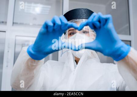 Femme médique dans un costume de protection et un masque en plastique fait des mains en forme de coeur Banque D'Images