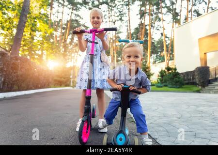 Deux adorables petits frères et sœurs caucasiens garçons en chemise et fille en robe ayant plaisir à monter trois roues équilibre vélo de course scooter dans la forêt du parc de la ville Banque D'Images