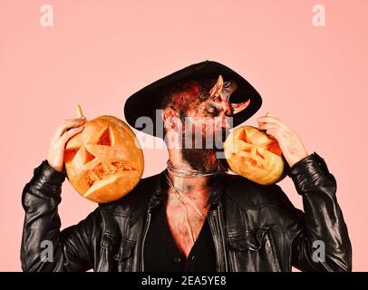 Homme portant un maquillage effrayant et des sorcières chapeau embrasse des citrouilles sur fond rose. Monstre avec décorations d'octobre. Démon avec des cornes et le visage occupé tient Jack o lanternes. Halloween – concept de fête effrayante Banque D'Images