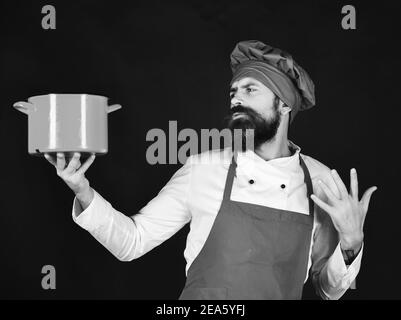 Concept du processus de cuisson. Chef avec une casserole ou une casserole rouge. Homme avec barbe tient les ustensiles de cuisine sur fond noir. Faites cuire avec un visage sérieux dans un chapeau bordeaux et le tablier tient le pot à soupe Banque D'Images