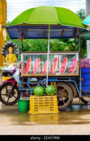 Chariot mobile de fruits ne vendant que des tranches de pastèque près d'un temple bouddhiste avec des statues de Bouddha en arrière-plan. Banque D'Images