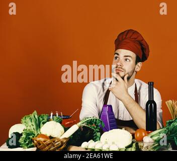 L'homme dans le chapeau de cuisinier et le tablier obtient une idée. Chef avec un visage bien pensé assis sur un fond rouge. Cuisiner travaille dans la cuisine près de la table avec des légumes et des outils. Ustensiles de cuisine et concept de cuisine. Banque D'Images