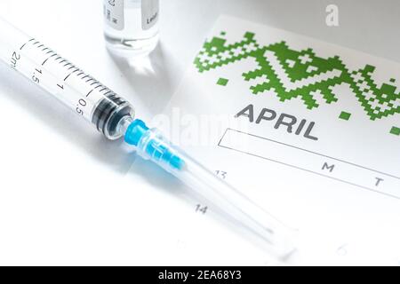 Gros plan d'une seringue médicale placée sur un calendrier d'avril avec un flacon de vaccin en arrière-plan, symbolisant la vaccination programmée Banque D'Images