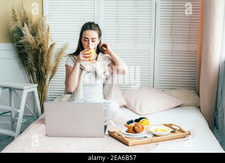 Jeune belle femme assise sur un lit prenant le petit déjeuner et communiquant dans un ordinateur portable. Apprentissage et communication à distance. Banque D'Images