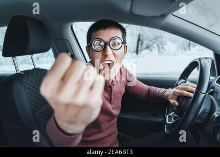 Drôle et ridicule conducteur nerd dans les lunettes criant à la caméra et montrant son poing à l'intérieur d'une voiture. Concept d'un conflit et d'un accID de conducteur novice Banque D'Images