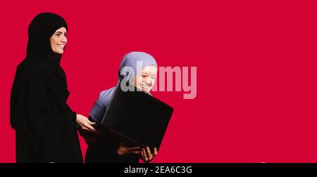 Bannière, format long. Une femme d'affaires musulmane dans le hijab riant heureusement avec un ordinateur portable pendant la journée de travail sur fond rouge avec espace publicitaire latéral. Banque D'Images