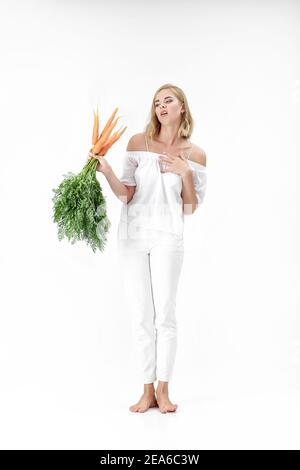 femme blonde dans un chemisier blanc tenant une carotte avec des feuilles vertes sur un fond blanc. fille se sent mauvais des carottes et des régimes alimentaires Banque D'Images